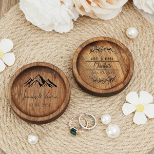 Peut inclure: Deux bo&icirc;tes &agrave; alliances en bois avec des motifs grav&eacute;s. Une bo&icirc;te repr&eacute;sente une cha&icirc;ne de montagnes avec le texte "Venus & Kevin 12.04.2024". L'autre bo&icirc;te a un motif floral avec le texte "mr & mrs Charlotte July 07. 2023".