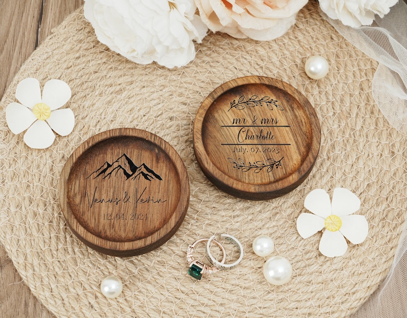 Peut inclure: Deux bo&icirc;tes &agrave; alliances en bois avec des motifs grav&eacute;s. Une bo&icirc;te repr&eacute;sente une cha&icirc;ne de montagnes avec le texte "Venus & Kevin 12.04.2024". L'autre bo&icirc;te a un motif floral avec le texte "mr & mrs Charlotte July 07. 2023".