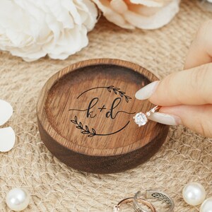 Peut inclure: Un porte-bague en bois avec une finition brun fonc&eacute;. Le plat a un design circulaire avec une bordure feuillue et les initiales "k+d" grav&eacute;es au centre. Une bague en diamant repose sur le plat.