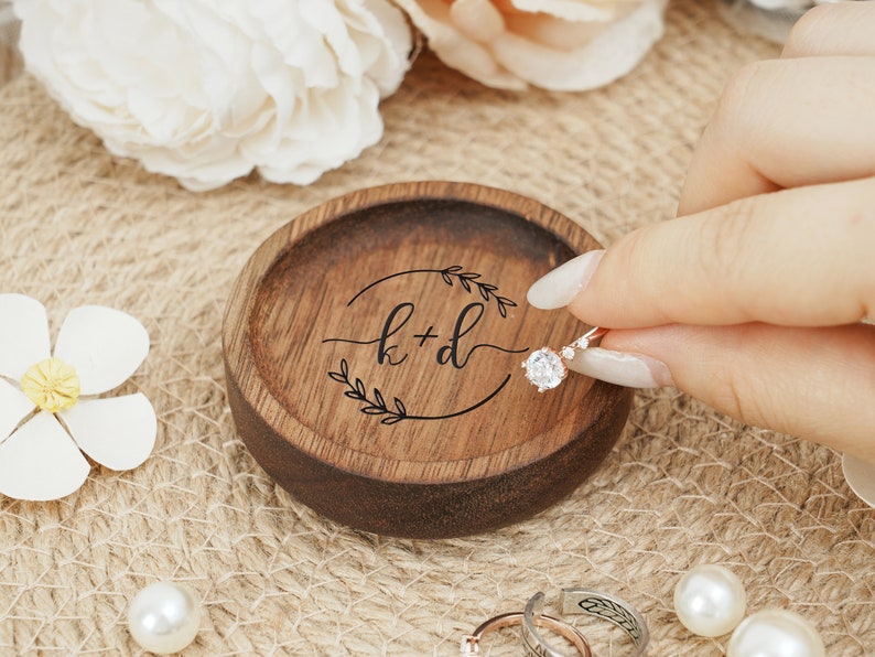 Peut inclure: Un porte-bague en bois avec une finition brun fonc&eacute;. Le plat a un design circulaire avec une bordure feuillue et les initiales "k+d" grav&eacute;es au centre. Une bague en diamant repose sur le plat.
