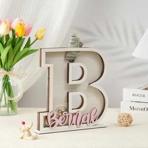 May include: White wooden letter B piggy bank with a slot for coins and bills. The letter B is decorated with a pink name "Bernal".