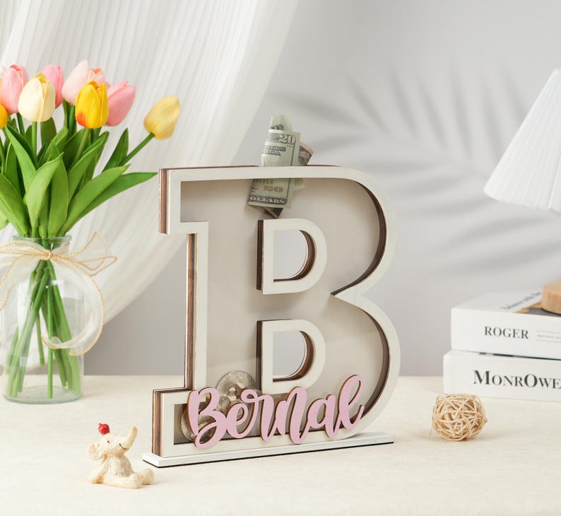 May include: White wooden letter B piggy bank with a slot for coins and bills. The letter B is decorated with a pink name "Bernal".