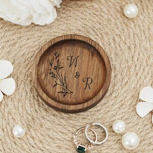 Peut inclure: Un porte-bagues en bois avec un motif floral et les initiales "W & R" grav&eacute;es sur le dessus. Le porte-bagues est pos&eacute; sur une surface tiss&eacute;e.