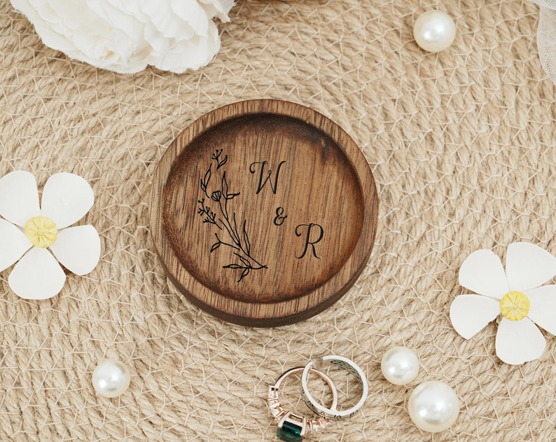 Peut inclure: Un porte-bagues en bois avec un motif floral et les initiales "W & R" grav&eacute;es sur le dessus. Le porte-bagues est pos&eacute; sur une surface tiss&eacute;e.