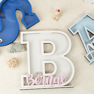 May include: White wooden letter B with a slot for coins. The letter is decorated with a pink name "Bernal" and stands on a white surface.