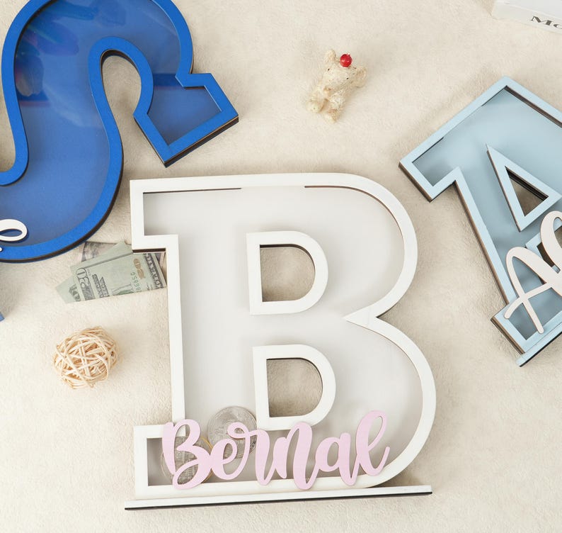 May include: White wooden letter B with a slot for coins. The letter is decorated with a pink name "Bernal" and stands on a white surface.