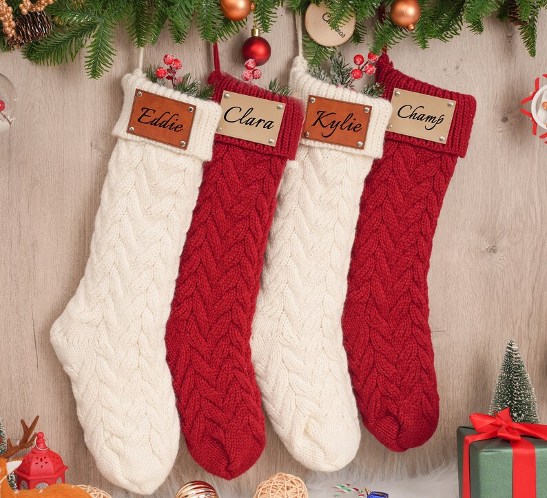 Op de afbeelding: Vier rode en witte gebreide kerstsokken met gepersonaliseerde houten naamschildjes. De namen op de naamschildjes zijn Eddie, Clara, Kylie en Champ.