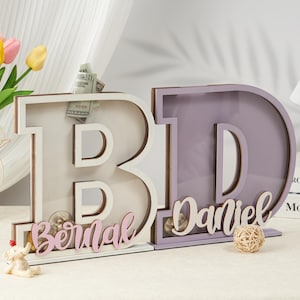 May include: Two wooden letter piggy banks, one white with the name "Bernal" in pink lettering and one purple with the name "Daniel" in white lettering.