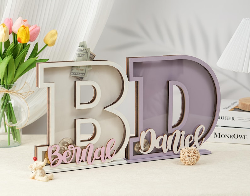 May include: Two wooden letter piggy banks, one white with the name "Bernal" in pink lettering and one purple with the name "Daniel" in white lettering.
