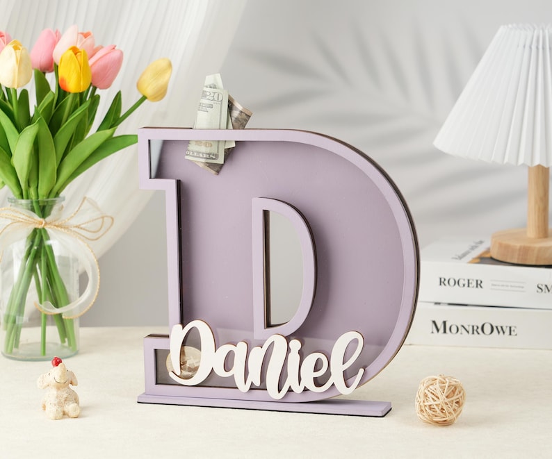May include: A light purple wooden letter D piggy bank with a slot for coins and a clear plastic window. The name "Daniel" is written in white on the front of the piggy bank.