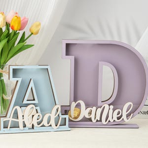 May include: Two wooden letter shaped boxes, one light blue and one light purple, with the names "Aled" and "Daniel" written on them. The boxes are sitting on a table with a bouquet of yellow and pink tulips in a glass vase.