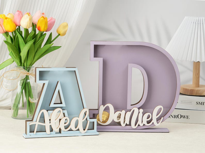 May include: Two wooden letter shaped boxes, one light blue and one light purple, with the names "Aled" and "Daniel" written on them. The boxes are sitting on a table with a bouquet of yellow and pink tulips in a glass vase.