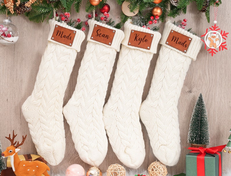 Op de afbeelding: Vier witte gebreide kerstsokken met bruine leren naamplaatjes. De namen op de naamplaatjes zijn Mads, Scan, Kylo en Mads.