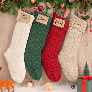 Op de afbeelding: Vier gebreide kerstsokken met gepersonaliseerde naamstickers. De sokken zijn cr&egrave;me, groen, rood en cr&egrave;me. De naamstickers zijn goudkleurig met zwarte letters. De namen op de stickers zijn Eric, Chase, Denal en Levon.