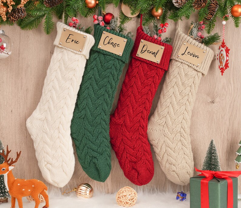 Op de afbeelding: Vier gebreide kerstsokken met gepersonaliseerde naamstickers. De sokken zijn cr&egrave;me, groen, rood en cr&egrave;me. De naamstickers zijn goudkleurig met zwarte letters. De namen op de stickers zijn Eric, Chase, Denal en Levon.