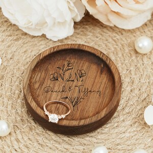 Peut inclure: Un porte-bague en bois avec un motif floral et les noms "David & Tiffany" grav&eacute;s dessus. Une bague en or repose dans le porte-bague.