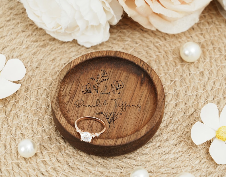 Peut inclure: Un porte-bague en bois avec un motif floral et les noms "David & Tiffany" grav&eacute;s dessus. Une bague en or repose dans le porte-bague.