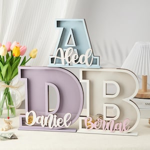 May include: Three wooden piggy banks shaped like letters, spelling out the name "Daniel Bernal". The first letter, "A", is light blue, the second letter, "D", is lavender, and the third letter, "B", is white. The name is spelled out in pink letters on each piggy bank.