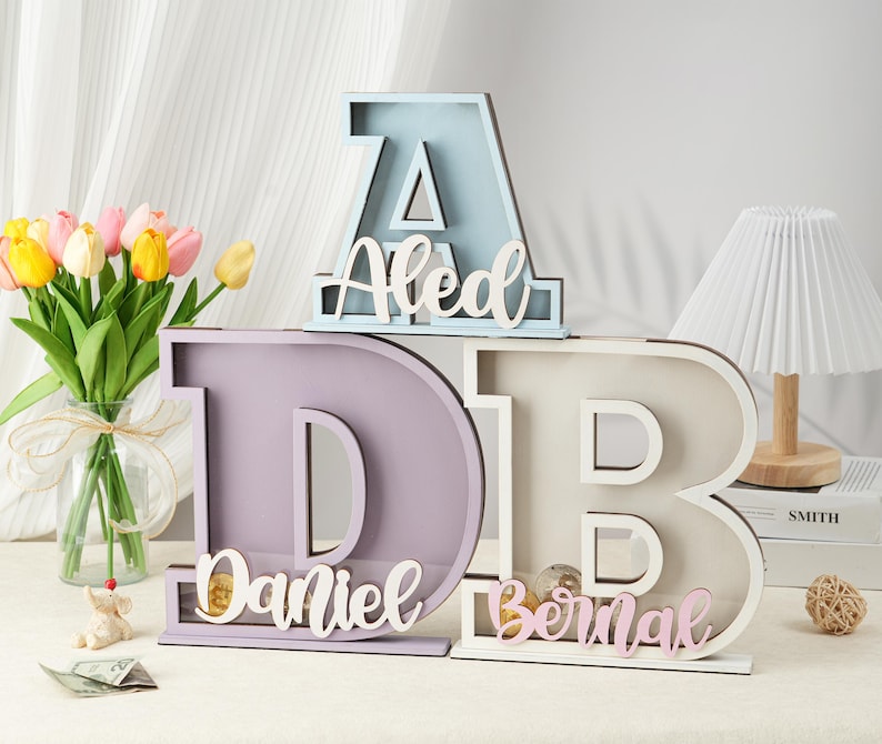 May include: Three wooden piggy banks shaped like letters, spelling out the name "Daniel Bernal". The first letter, "A", is light blue, the second letter, "D", is lavender, and the third letter, "B", is white. The name is spelled out in pink letters on each piggy bank.