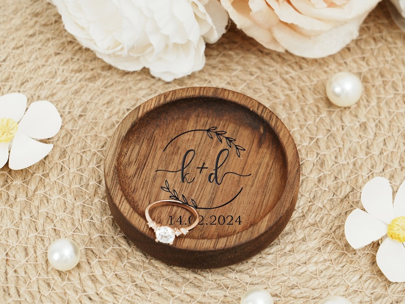 Peut inclure: Un porte-bague en bois avec une finition brun fonc&eacute;. Le plat a un cercle grav&eacute; avec les initiales "k+d" et la date "14.02.2024". Une bague en or repose sur le plat.