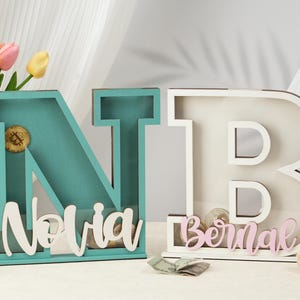May include: Two wooden letter piggy banks, one blue and one white, with the names "Novia" and "Bernal" painted on them. The letters are standing on a table with a bouquet of flowers and a lamp in the background.