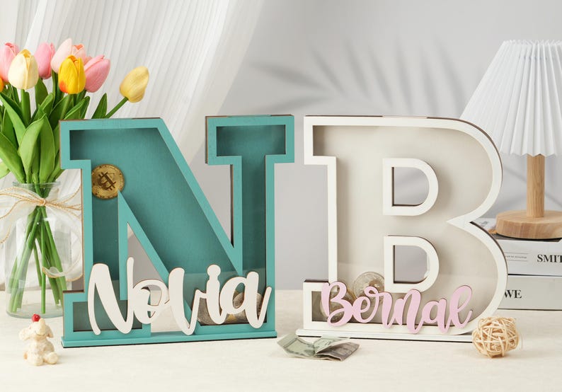 May include: Two wooden letter piggy banks, one blue and one white, with the names "Novia" and "Bernal" painted on them. The letters are standing on a table with a bouquet of flowers and a lamp in the background.