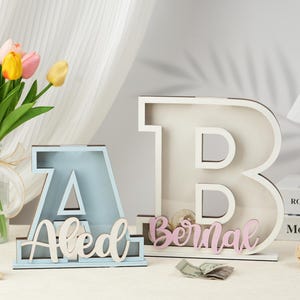 May include: Two wooden letter piggy banks, one blue and one white, with the names "Aled" and "Bernal" spelled out in white and pink letters. The piggy banks are on a white surface with a bouquet of tulips in the background.