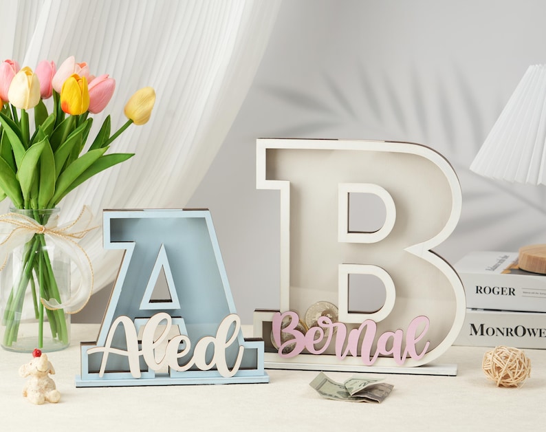 May include: Two wooden letter piggy banks, one blue and one white, with the names "Aled" and "Bernal" spelled out in white and pink letters. The piggy banks are on a white surface with a bouquet of tulips in the background.