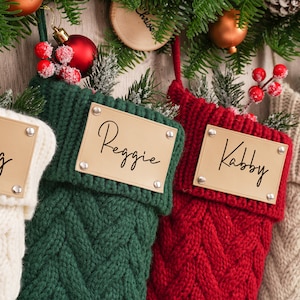 Op de afbeelding: Vier gebreide kerstsokken met gepersonaliseerde naamstickers. De sokken zijn wit, groen, rood en beige. De namen op de stickers zijn Craig, Reggie, Kabby en Jake.