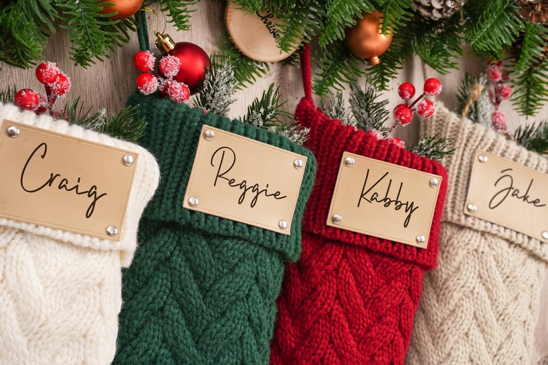 Op de afbeelding: Vier gebreide kerstsokken met gepersonaliseerde naamstickers. De sokken zijn wit, groen, rood en beige. De namen op de stickers zijn Craig, Reggie, Kabby en Jake.