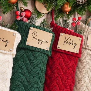 Könnte beinhalten: Vier Zopfstrick-Weihnachtsstrümpfe in Creme, Grün, Rot und Beige, jeweils mit personalisiertem Namensschild. Sie sind mit festlichem Grün, Beeren und Ornamenten geschmückt, was ein festliches Thema ergibt.