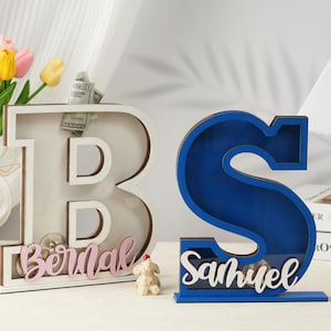 May include: Two wooden piggy banks shaped like the letters B and S. The B is white with pink lettering and the S is blue with white lettering. The B has the name "Bernal" on it and the S has the name "Samuel" on it.