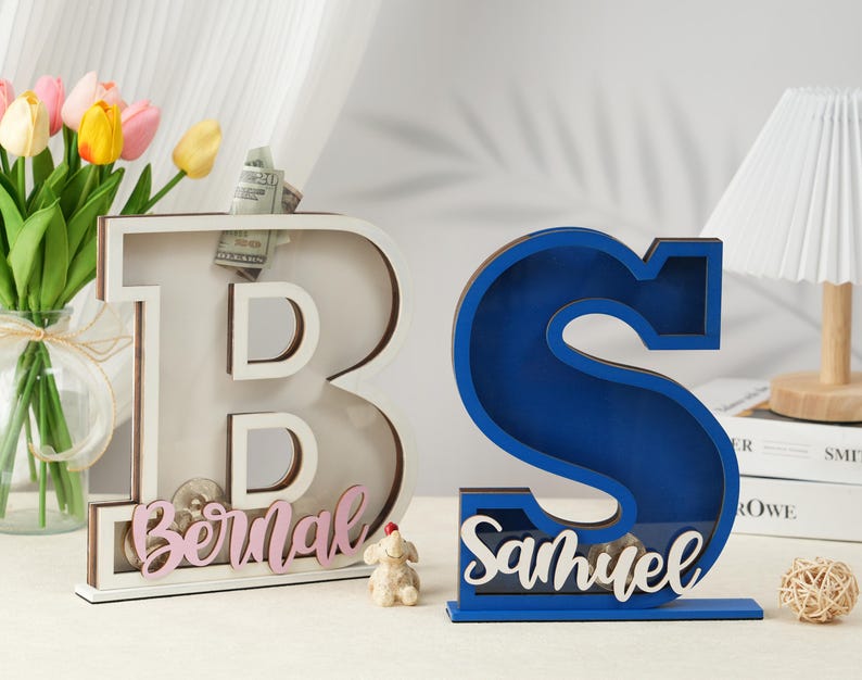 May include: Two wooden piggy banks shaped like the letters B and S. The B is white with pink lettering and the S is blue with white lettering. The B has the name "Bernal" on it and the S has the name "Samuel" on it.