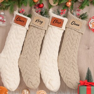 Op de afbeelding: Vier beige en witte gebreide kerstsokken met gepersonaliseerde leren naamstickers. De namen op de stickers zijn Dave, Rory, Elize en Daan.