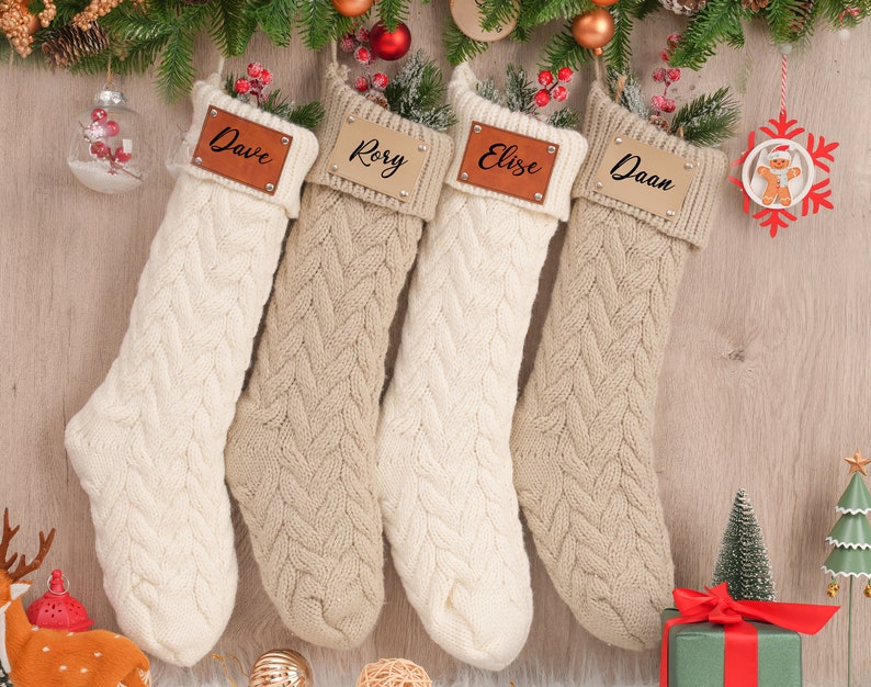 Op de afbeelding: Vier beige en witte gebreide kerstsokken met gepersonaliseerde leren naamstickers. De namen op de stickers zijn Dave, Rory, Elize en Daan.