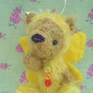 Pode incluir: Um urso de peluche amarelo e peludo vestindo um traje de borboleta amarelo com botões laranja e vermelho. O urso tem um nariz bordado preto e olhos pretos.