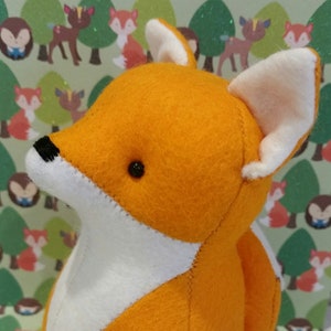 May include: A plush orange fox toy with white ears and a white belly. The fox is sitting on a green background with a pattern of deer and foxes.