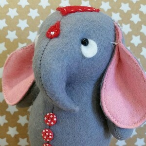 Può includere: Un giocattolo di elefante in feltro grigio con orecchie rosa e dettagli rossi. L'elefante indossa un cappello rosso e ha tre bottoni rossi con pois bianchi sul suo corpo. L'elefante è in piedi su una palla bianca e rossa.
