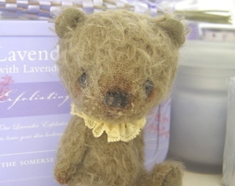 May include: A small, brown teddy bear with a yellow lace collar. The bear is sitting on a white surface. The bear has a fuzzy brown coat and a sweet expression.
