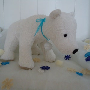 May include: A plush white polar bear toy with a blue ribbon and bell around its neck. The bear is surrounded by artificial snow and snowflake decorations. The bear has black eyes and a black nose.