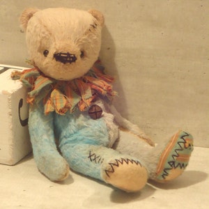 May include: A handmade plush teddy bear with a light brown fur, blue and white clothing, and a colorful patterned collar. The bear has a button nose and is sitting on a white surface.