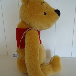 May include: A plush Winnie the Pooh bear wearing a red vest. The bear is sitting with its right leg extended and its left leg tucked under its body. The bear has a tag on its left leg that reads "Good Bears by Maessen".