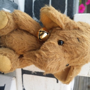 May include: A brown plush elephant toy with a gold bell around its neck. The elephant has a soft, fuzzy texture and large, dark eyes.