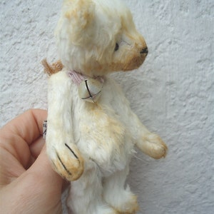 May include: A white teddy bear with a pink ribbon and a silver bell around its neck. The bear is standing on a white surface.