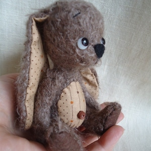 May include: A brown and tan plush bunny rabbit with a button nose and brown eyes. The bunny is wearing a brown and tan patterned fabric on its ears and chest.