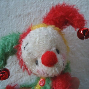 May include: A white plush toy with a red nose and yellow, green, and red fur. The toy has two red bells attached to its ears.