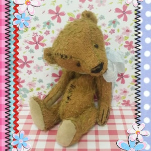 May include: A 4.5" Buddy mc Bear pattern for making a plush toy. The pattern is shown with a finished brown teddy bear with a white bow sitting on a pink and white checkered surface. The background is a pink and white floral print.
