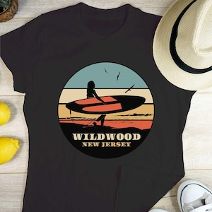 Puede incluir: Camiseta negra con un dise&ntilde;o gr&aacute;fico circular que representa a un surfista en una tabla de surf. El dise&ntilde;o muestra una escena de playa con cielo azul, puesta de sol naranja y roja, y el texto "Wildwood New Jersey". Se ve un sombrero de paja.