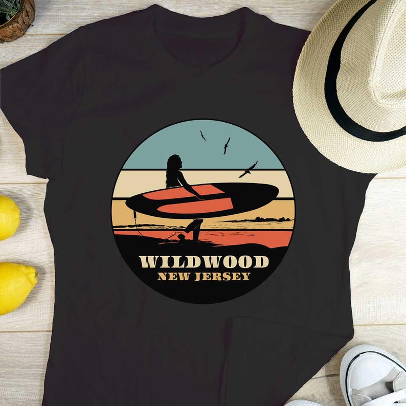 Puede incluir: Camiseta negra con un dise&ntilde;o gr&aacute;fico circular que representa a un surfista en una tabla de surf. El dise&ntilde;o muestra una escena de playa con cielo azul, puesta de sol naranja y roja, y el texto "Wildwood New Jersey". Se ve un sombrero de paja.