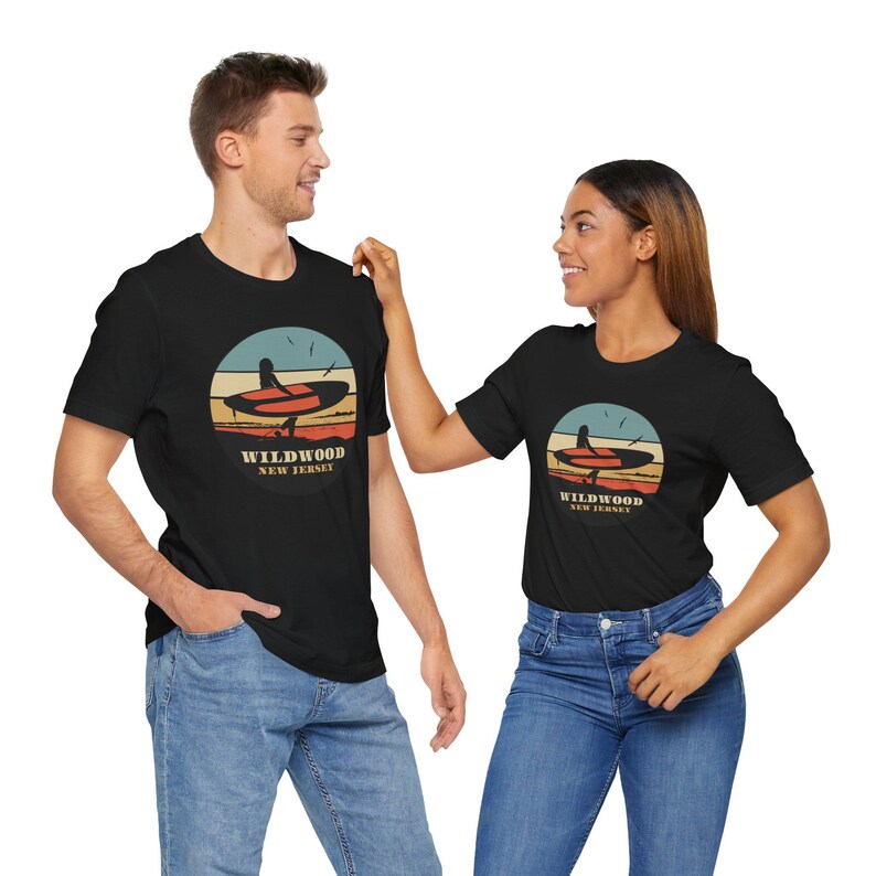 Puede incluir: Camisetas negras con un dise&ntilde;o gr&aacute;fico circular que muestra una tabla de surf, una puesta de sol y el texto "Wildwood New Jersey". Un hombre y una mujer llevan las camisetas con vaqueros azules.